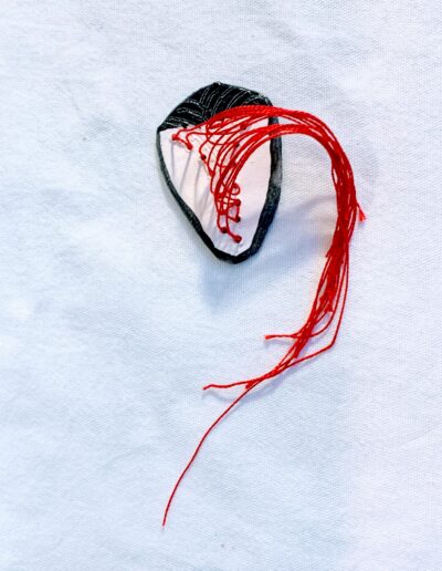 Forme blanche aux contours arrondis, légèrement triangulaire et terminée en pointe, avec une partie noire en relief et striée. De nombreux fils rouges la traversent et se prolongent librement vers le bas.